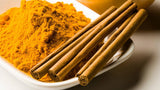 Baltame inde yra ryškiai oranžinis Prieskonių mišinys "GOLDEN DRINK", 120g Natural Market, šalia trijų sveikų cinamono lazdelių, besiremiančių ant jo krašto. Šalia jų gyva žaluma suteikia nedidelis Matcha miltelių piliakalnis. Fonas švelniai apšviestas, išryškinantis šiltus atspalvius ir kontrastingas prieskonių bei arbatos spalvas.